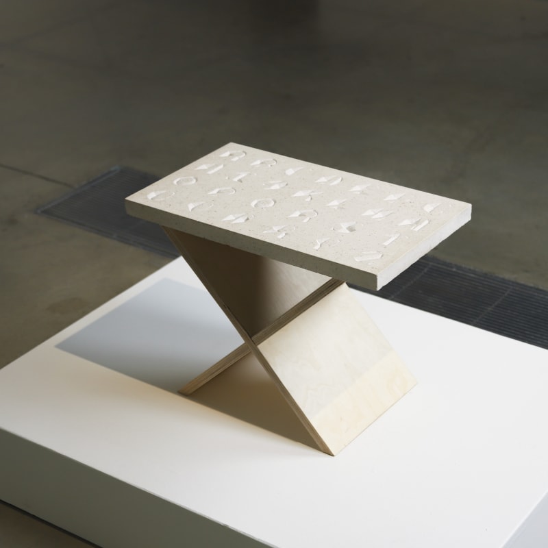 A sans serif typeface inspired by tombstone lettering hand carved in Portland limestone on a pale slab, mounted on an exhibition stand and shot from above and to the side.