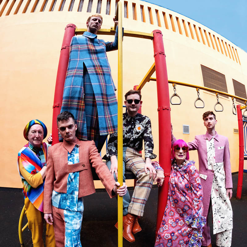 A group of six people, each wearing bold, colourful, and eclectic outfits, stands and sits on playground equipment against a brightly painted yellow wall.