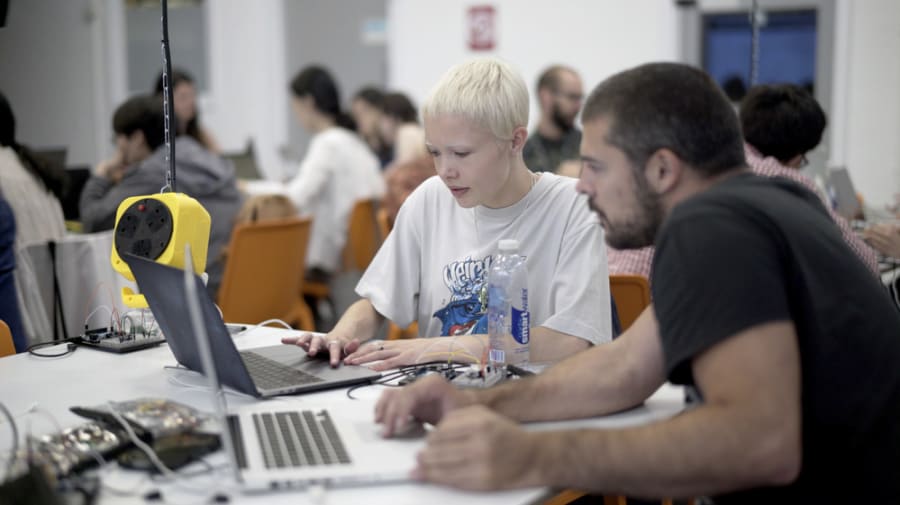 2 students working on laptops in the foreground. In the background there are other students working at other tables. 