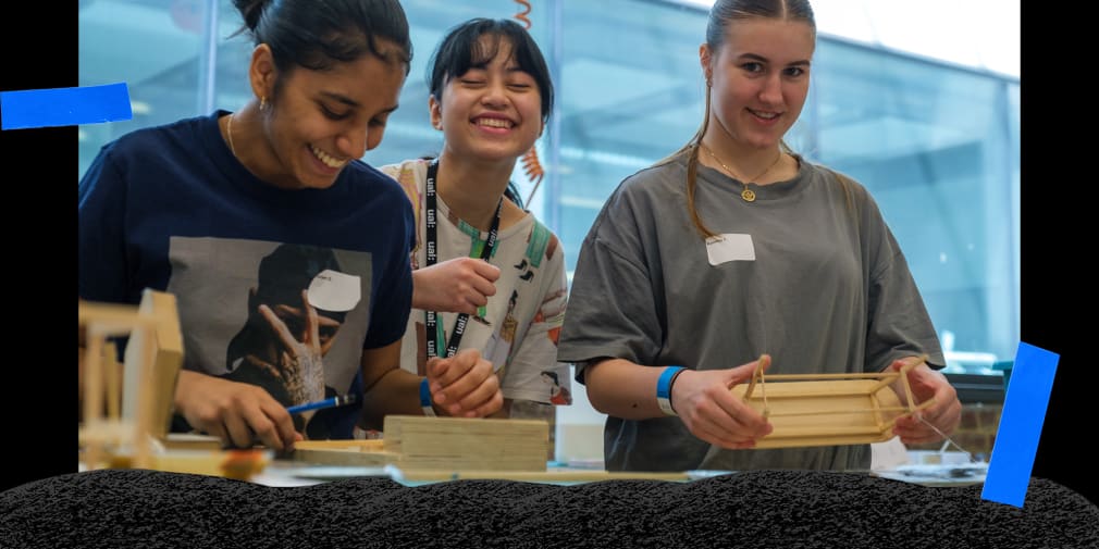 A group of students standing together in the technical workshop smiling and looking at the camera. 1 student is holding a scale models of a piece of furniture