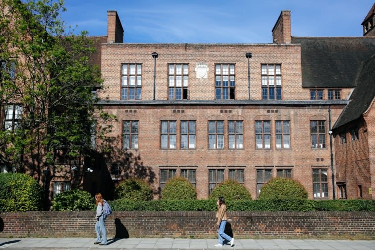 Two people walking on the street in front of an grand, old brick building.