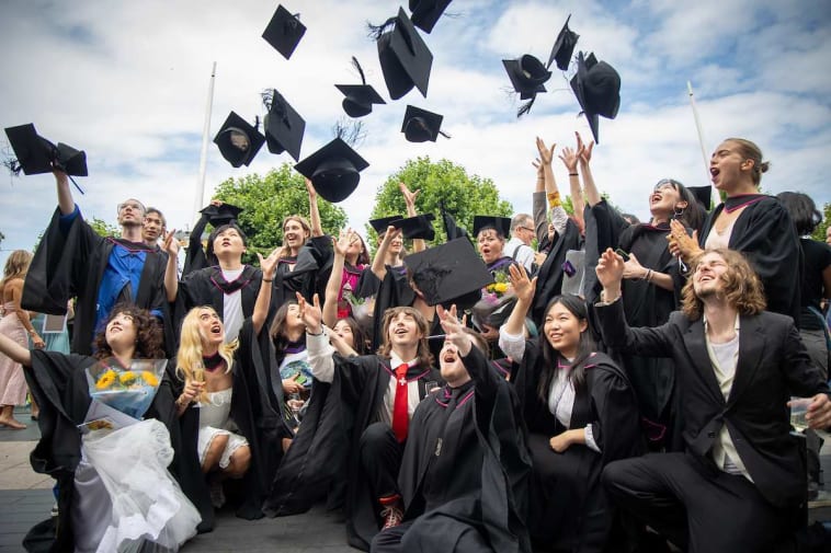 A group of graduates dressed in black gowns and caps celebrates outdoors, joyfully tossing their caps into the air. They are surrounded by greenery under a bright blue sky, smiling and cheering in celebration.
