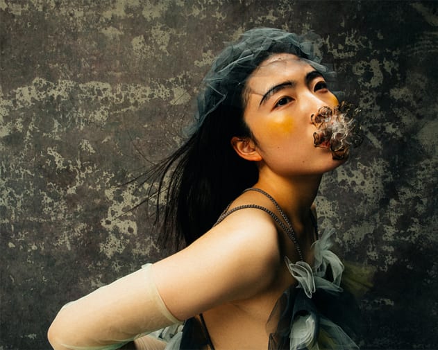 Model wearing green mesh sleeves, green head dress and artefact in her mouth.