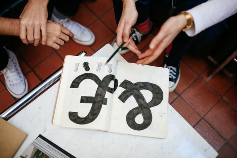 Close-up of 2 people's hands gesturing at a notepad on a table. 