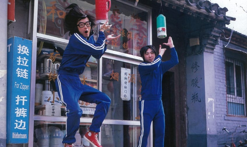 Two men in blue striped tracksuits jumping in the air in front of an Asian shop.