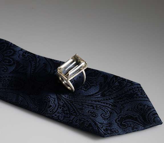A silver ring with a baguette or rectangular style glass stone is placed on a textured navy tie