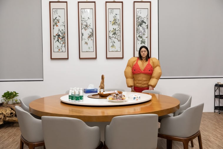 Image of woman wearing an inflatable superwoman suit standing in a formal meeting room setting. 