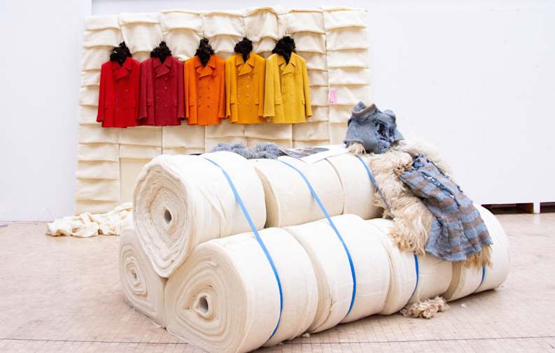 Bales of wool tied in blue bands, with knitted garments laid on top. In the background are pleated columns of wool with identical hanging tweed jackets in shades of red, orange and yellow.