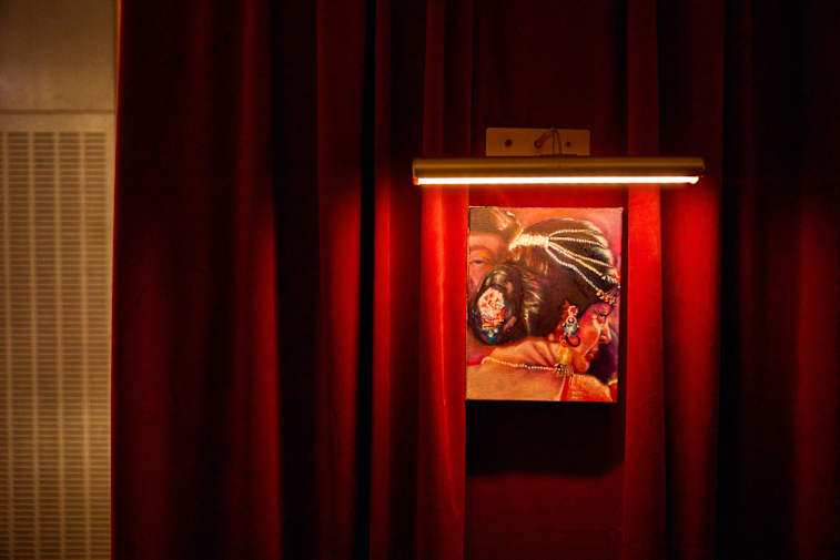 Red velvet curtains are slightly open, showing part of a painting that looks like a film still. Two characters are embracing and we just see the hair and eyes of one and the back of the other's head, with dark hair in a bun and jewelled hairpiece, necklace, earrings and other accessories.