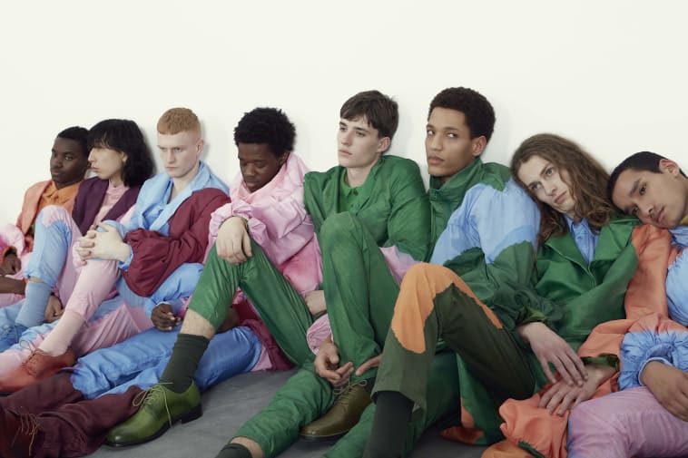 Male models in colourful outfits, standing together backstage.