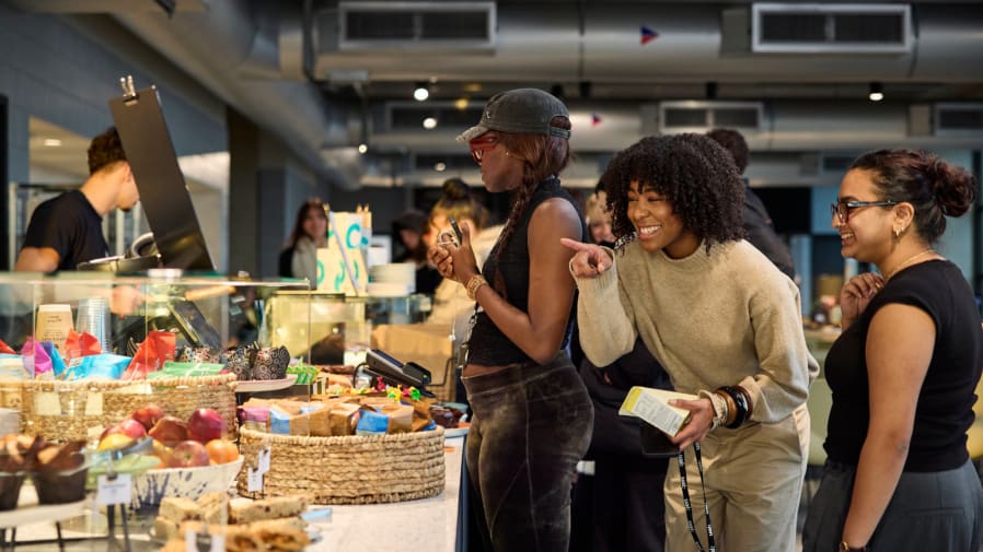 Group of diverse young adults interacting cheerfully while selecting snacks at a modern self-service café counter. The display features baskets filled with colorful packaged treats, fresh apples, muffins, and granola bars. One woman in a beige sweater enthusiastically points at an item, while others smile and look on, creating a vibrant atmosphere of community and casual social engagement. Industrial-style ceiling ducts and modern lighting enhance the urban café aesthetic.