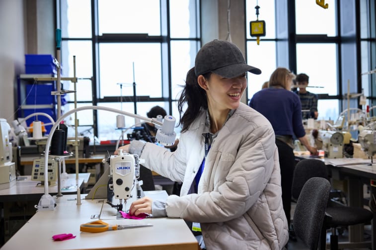 A person wearing a quilted jacket and a baseball cap sits at a sewing workstation in a bright, spacious studio. The person is operating an industrial sewing machine with a piece of bright pink fabric under the needle. The workstation includes thread spools, scissors, and various sewing tools. Several other people are working at sewing machines in the background. Large windows fill the room with natural light, and shelves and equipment are visible throughout the studio.