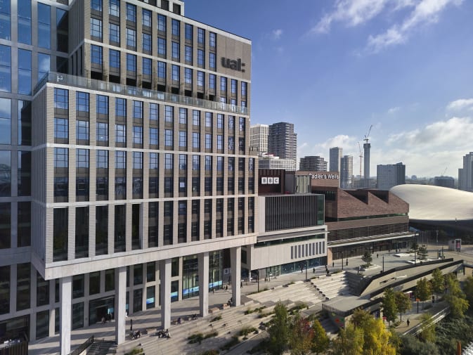 The LCF East Bank campus and building, shot from a side angle against a blue sky, with Sadler's Wells and BBC buildings and signage visible beside LCF.