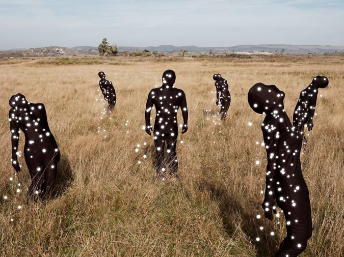 Several human figures in full black bodysuits stand and lean in a grassy landscape, their bodies covered with glowing white dots, against a backdrop of hills and open sky.