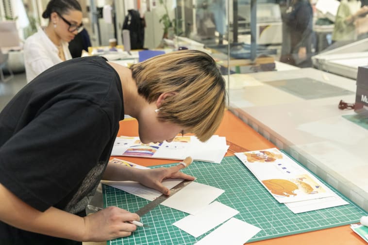 Person in a black top leaning over a green, gridded cutting mat and trimming small white cards
