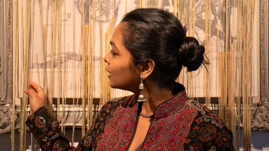 One Indian student stands in front of her artwork which hangs on the wall behind. She is turning to her right as she holds part of the work on her multidimensional canvas piece.