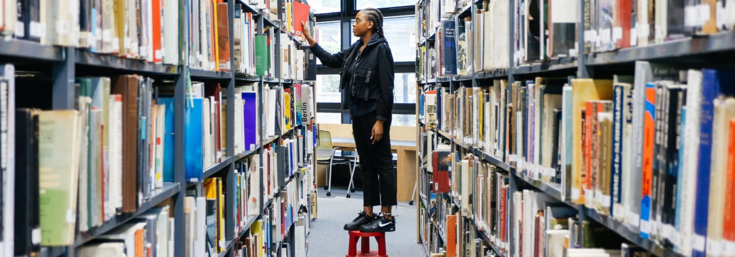 Student standing on a stool in the middle of two bookshelves.