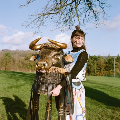Female in fireld with woodland behind, holding a large helmet of a horned mammal's head.