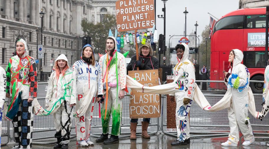 People wearing white boilersuits are connected to eachother, while a man holds a placard calling to ban plastic pollution