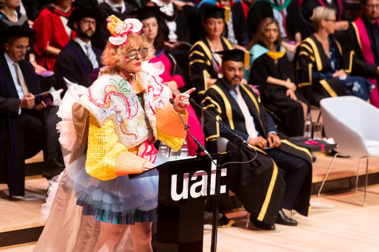Photo showing Sir Grayson Perry wearing Yuze Li's winning graduation gown and details of the gowns, on stage at Royal Festival Hall, as he addresses graduates.
