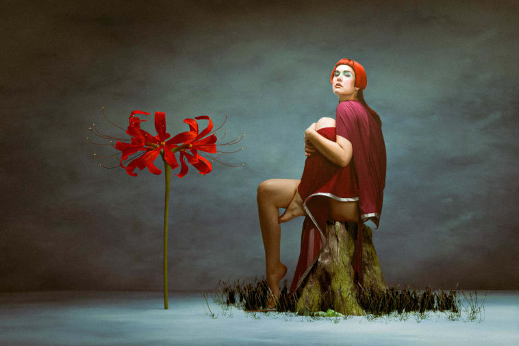 Model with bright red hair sitting on tree stump next do life size red flower.