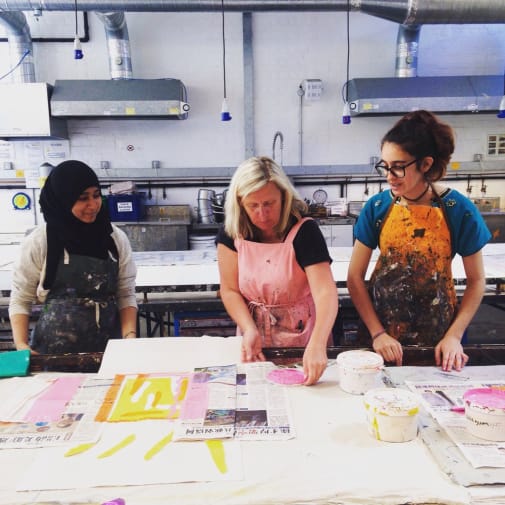 Printmaking tutor in centre of image with a student either side.  All 3 wearing ink spattered aprons whilst tutor holds paper over newsprint covered bench.