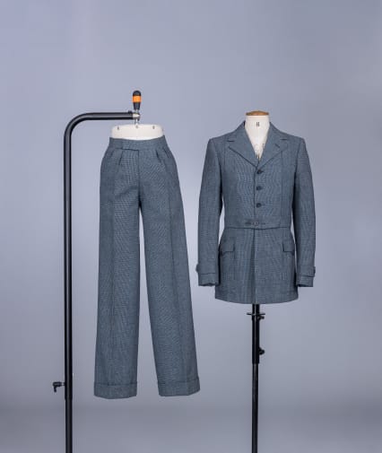 A grey two piece suit, on separate cream bodice and legs mannequins, supported by black metal stands, on a white backdrop