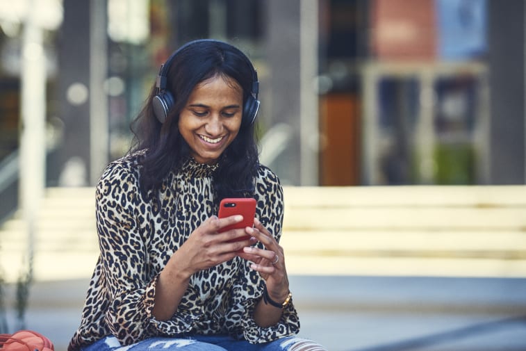 A smiling woman looking at her phone. She has long black hair and is wearing a pair of over ear headphone. 