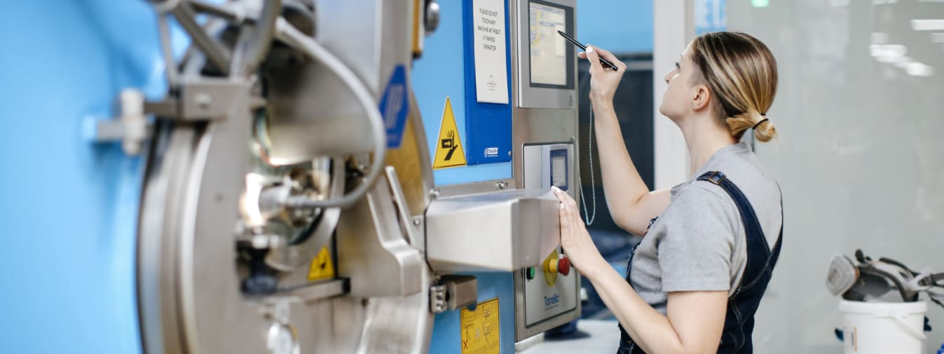 Leanne, a white woman with blonde hair, operating a Tonello denim washing machine, a large blue and grey machine with a series of yellow warning stickers on it 
