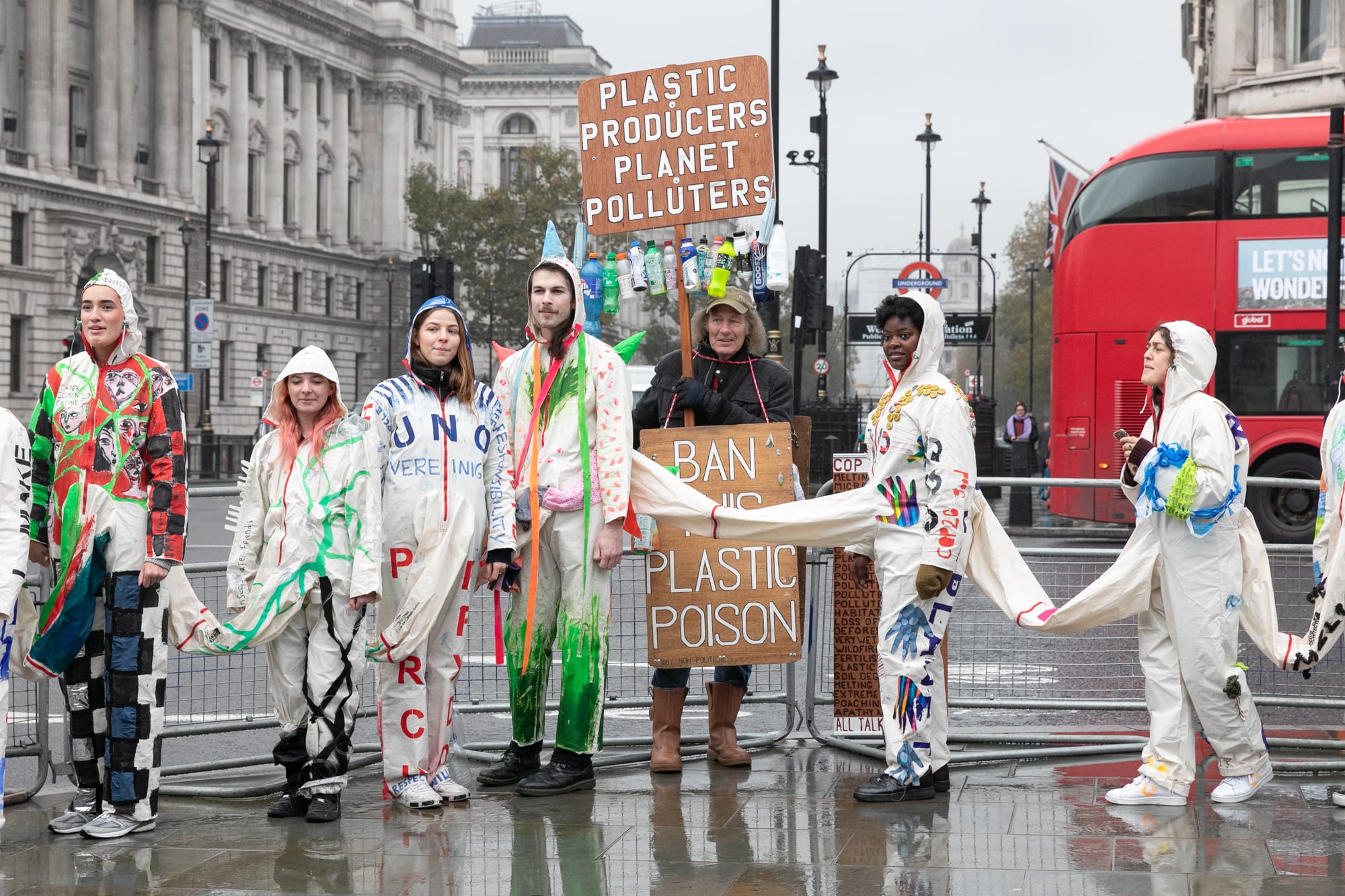  Image of students in Central London stood on the pavement wearing the 50-person conjoined garment with people holdings protest placards. You can see traffic of buses and cars in the background.