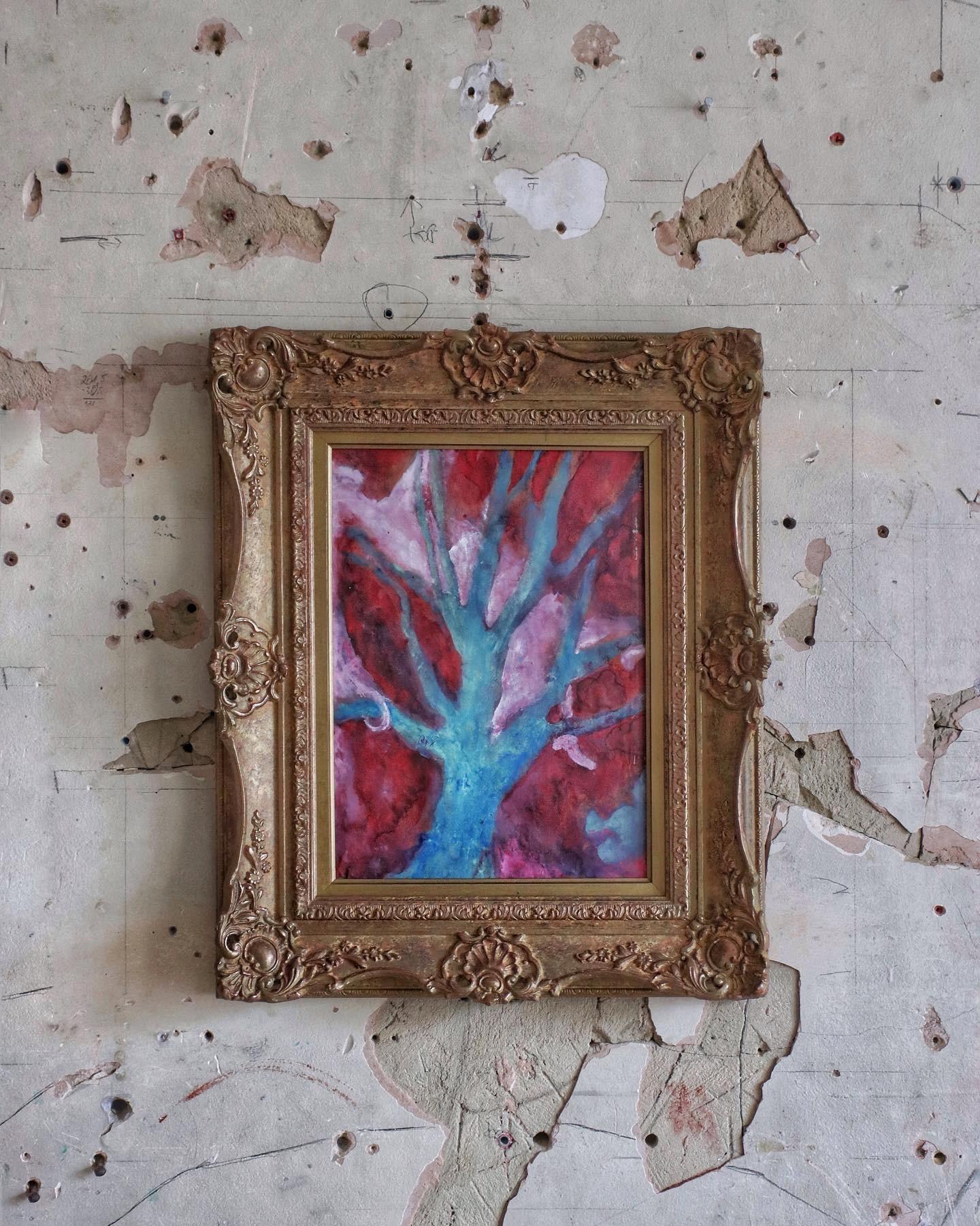 A small painting in blue and pink, with an ornate gold frame on a wall with peeling white paint