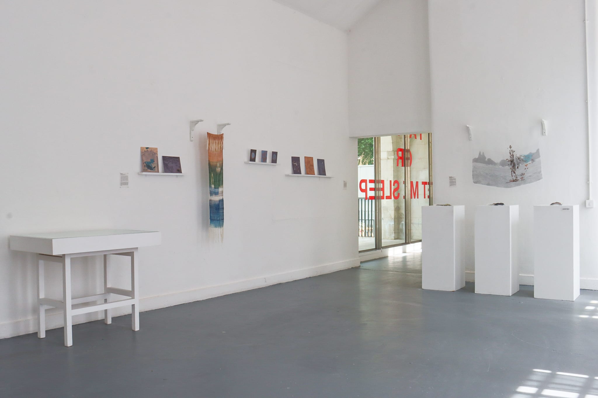 Art gallery interior with white walls and gray floor, exhibiting small artworks on shelves, and three white pedestals with a window displaying text.