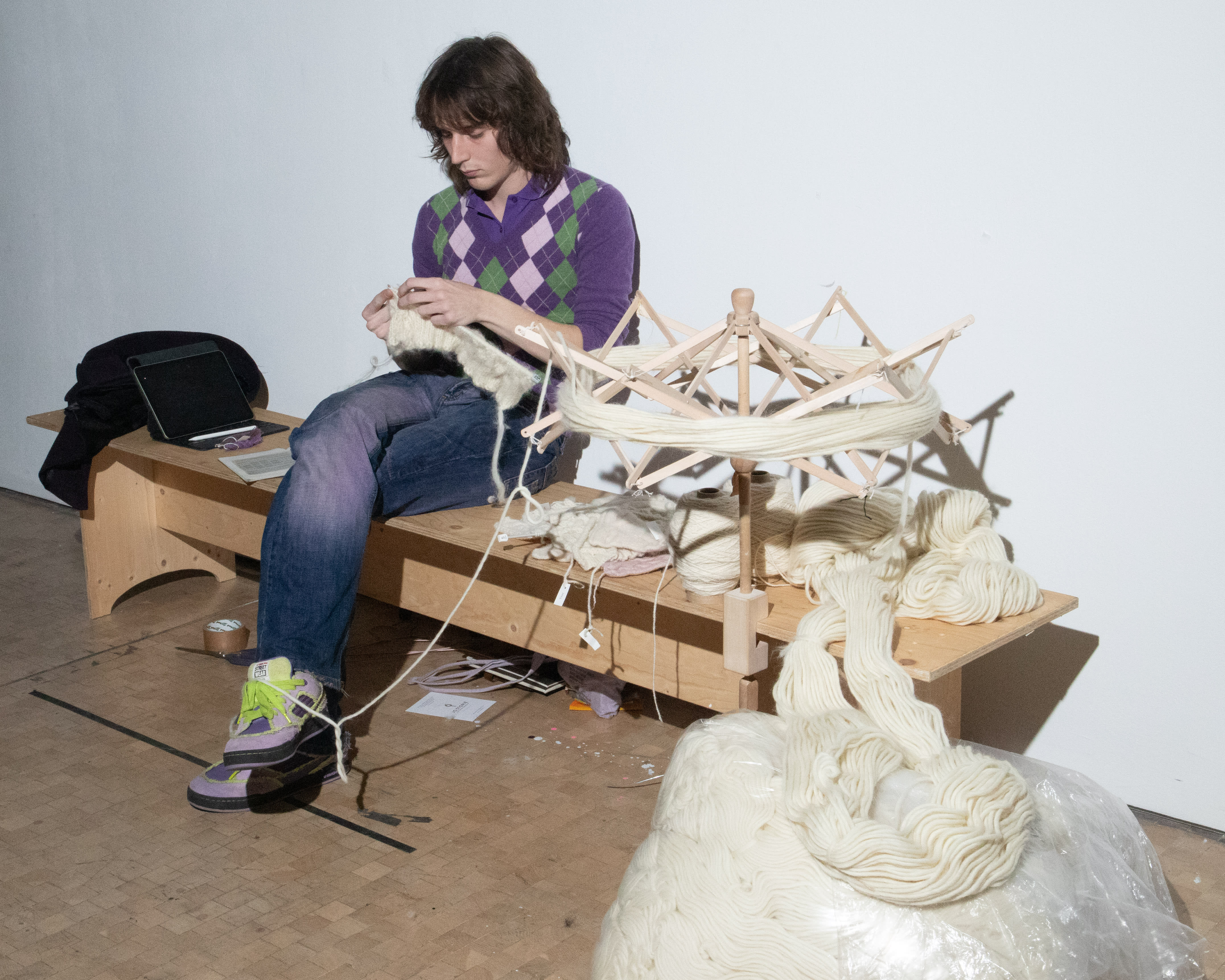 A student wearing an argyle pattern sweater and jeans is sitting with knees crossed working with a wool construction, next to a large bag of wool.