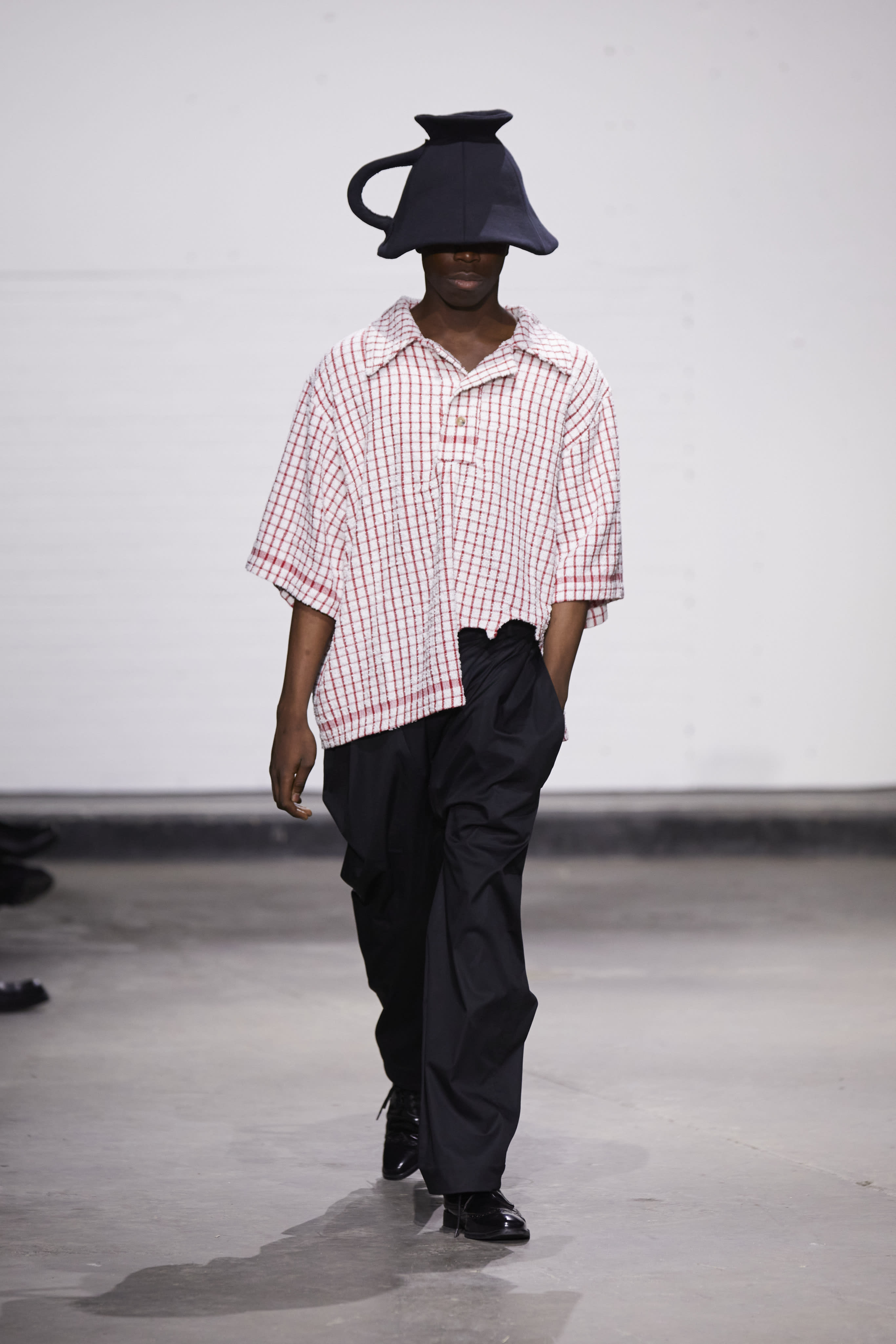 A model wearing a black upsidedown teapot shaped hat, a slouchy patterned shirt and dark loose trousers.