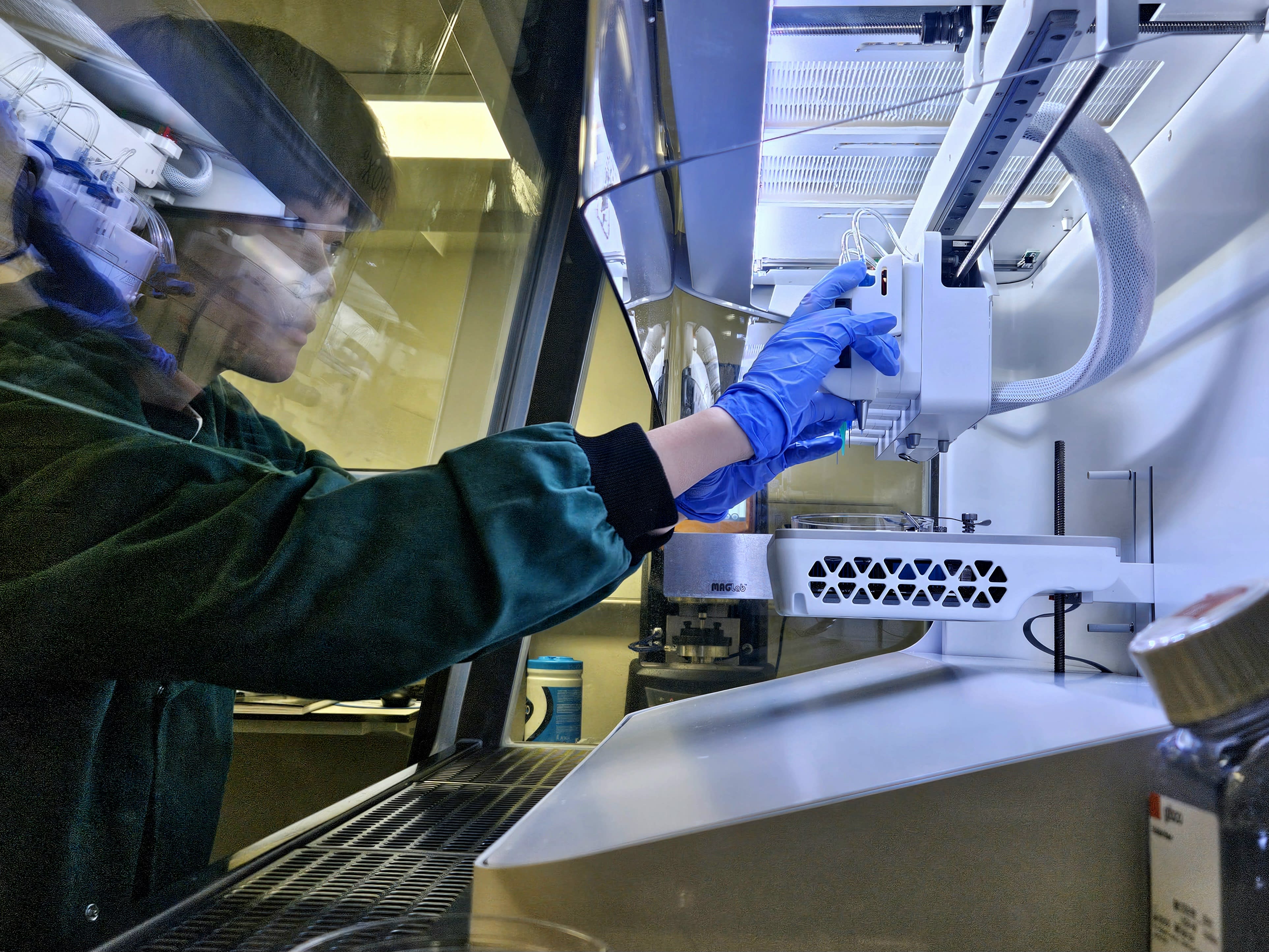 A person working with machinery in a lab. They are wearing a white coat and safety glasses.