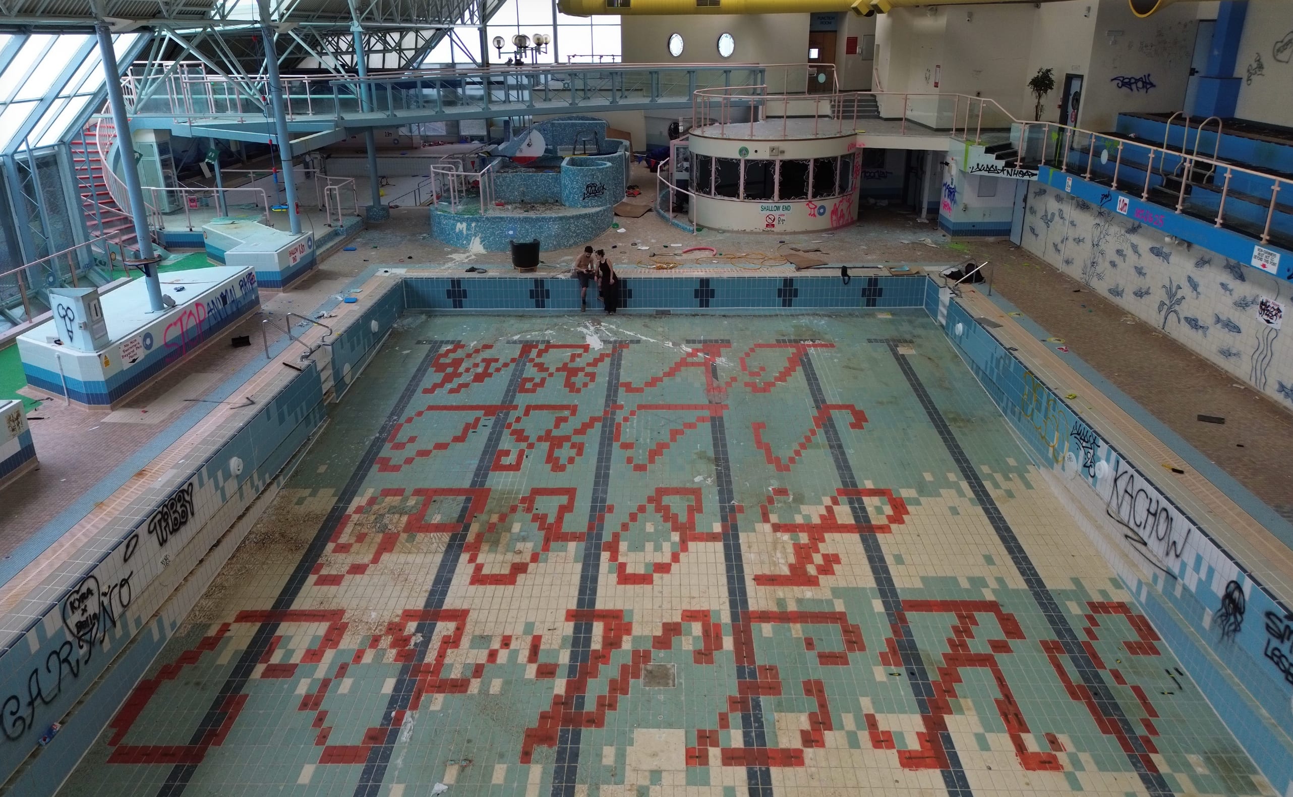 An empty indoor pool, with blue and whitetiled floor and walls and red decorative pattern that looks like lettering; there is also graffiti and industrial-looking equipment and discarded materials including an armchair. Two young people are having a close conversation at the far end. 