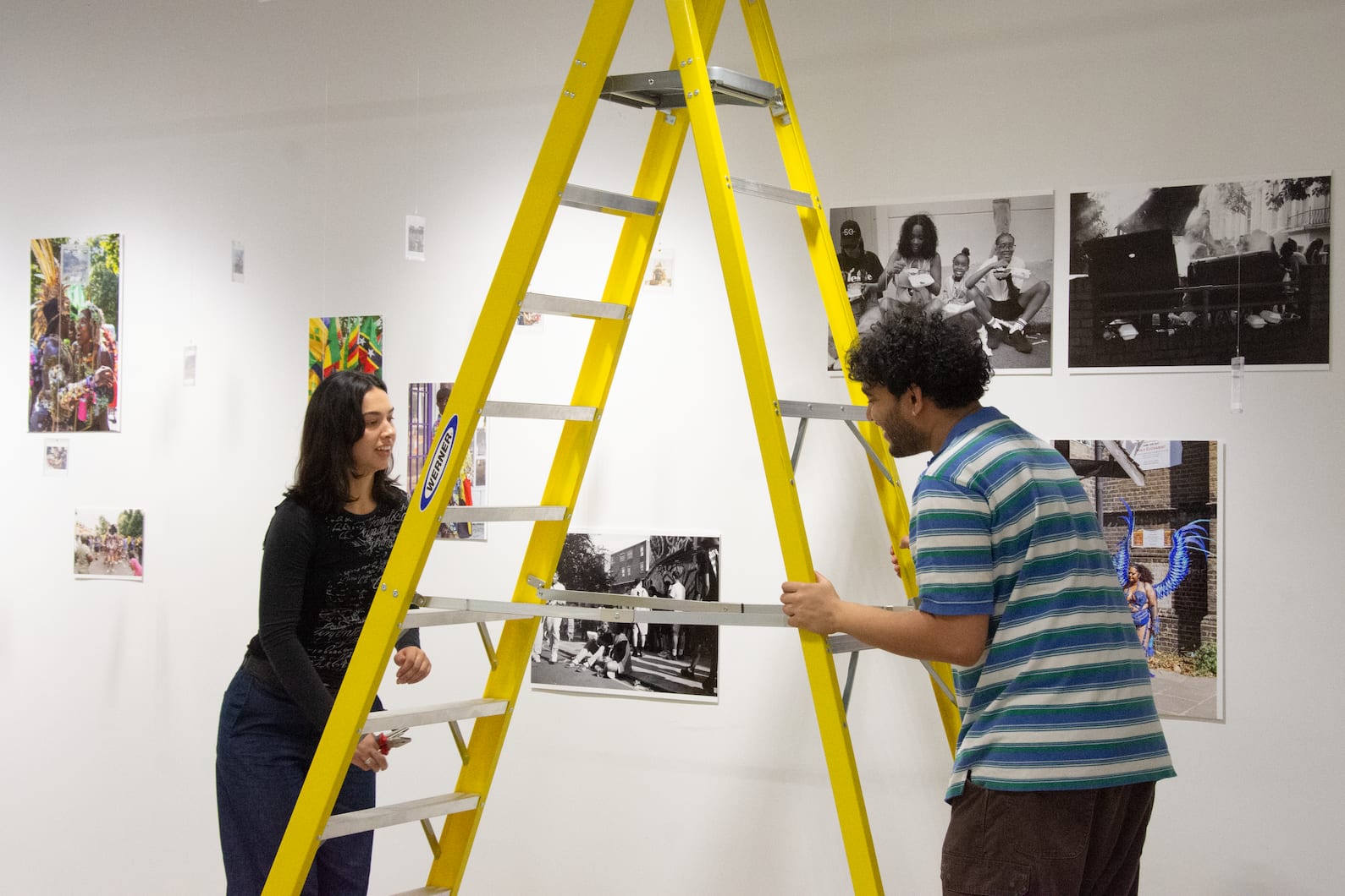 Two students holding either side of a yellow stepladder as they install printed photographic works on to white walls