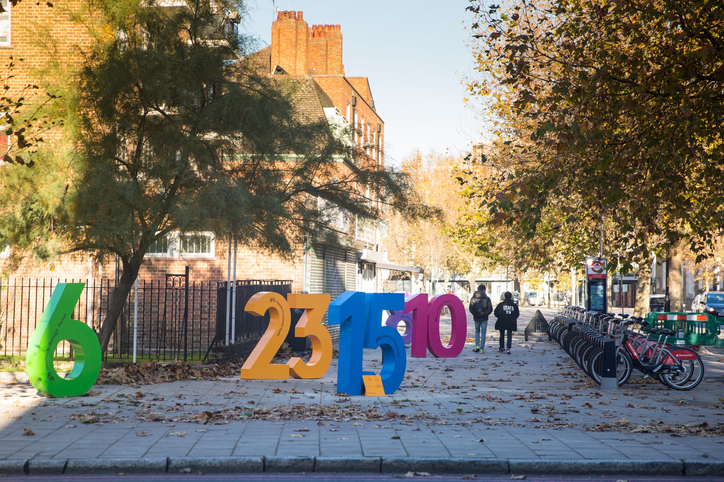 Large colourful numbers standing on a street by a bike hire bay.