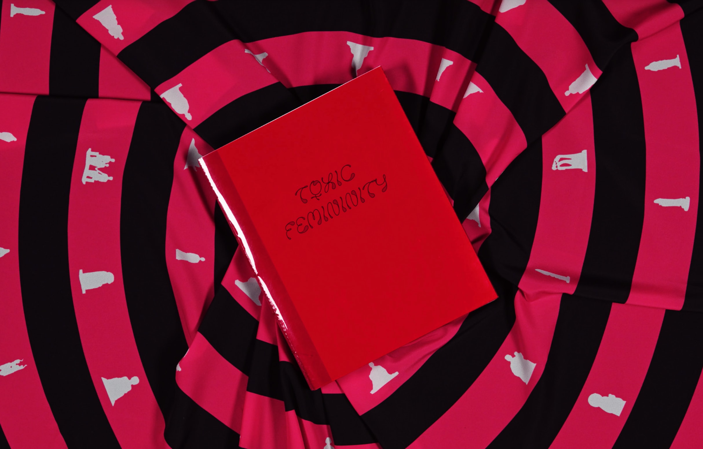 A book with a red cover and decorative text title TOXIC FEMININITY. It is at the centre of what looks like a spiral patterned fabric in red and black with white symbols and figures at intervals.