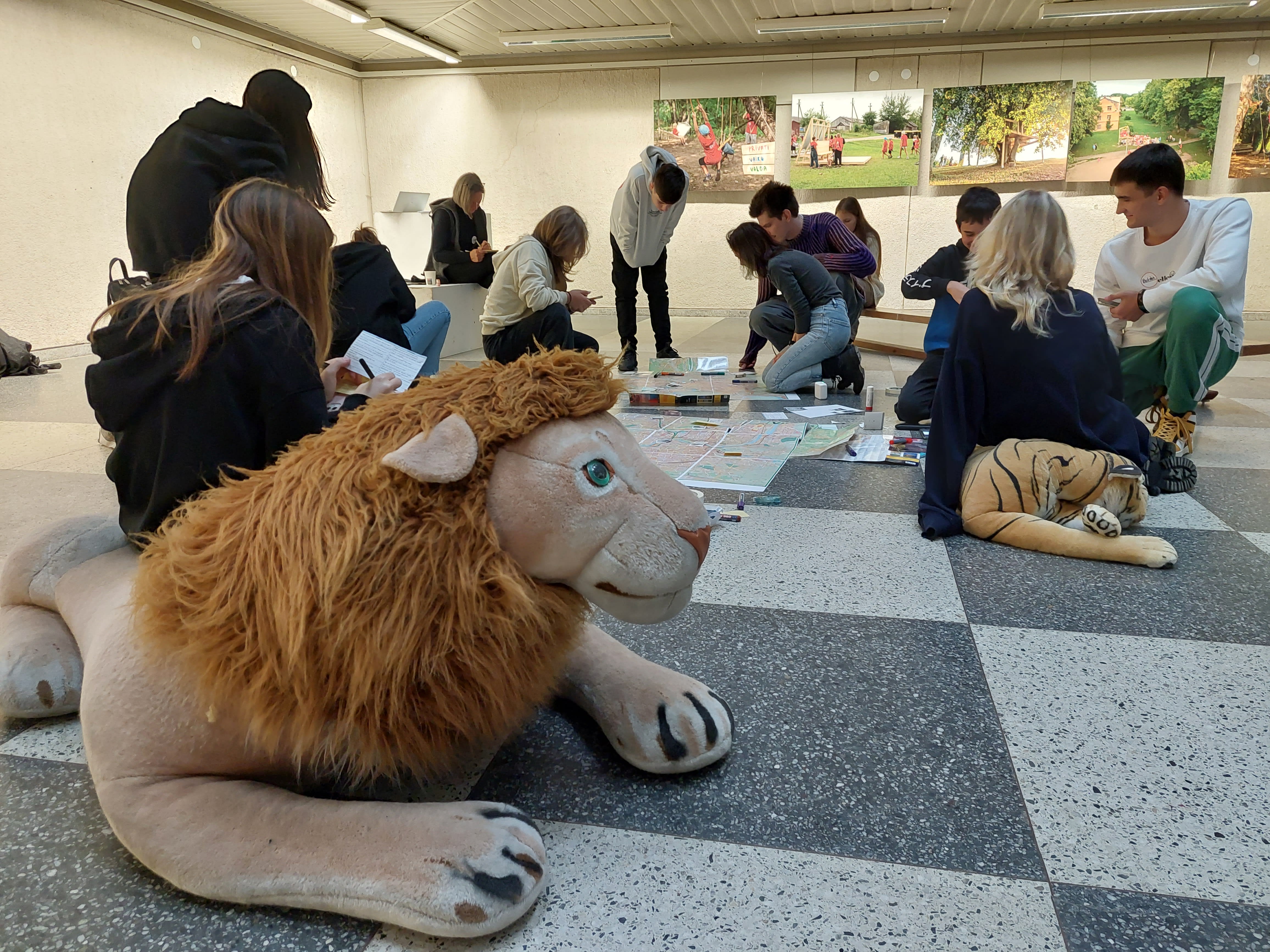 Teenagers and project team seated and kneeling on a square tiled floor with Simba, the stuffed lion