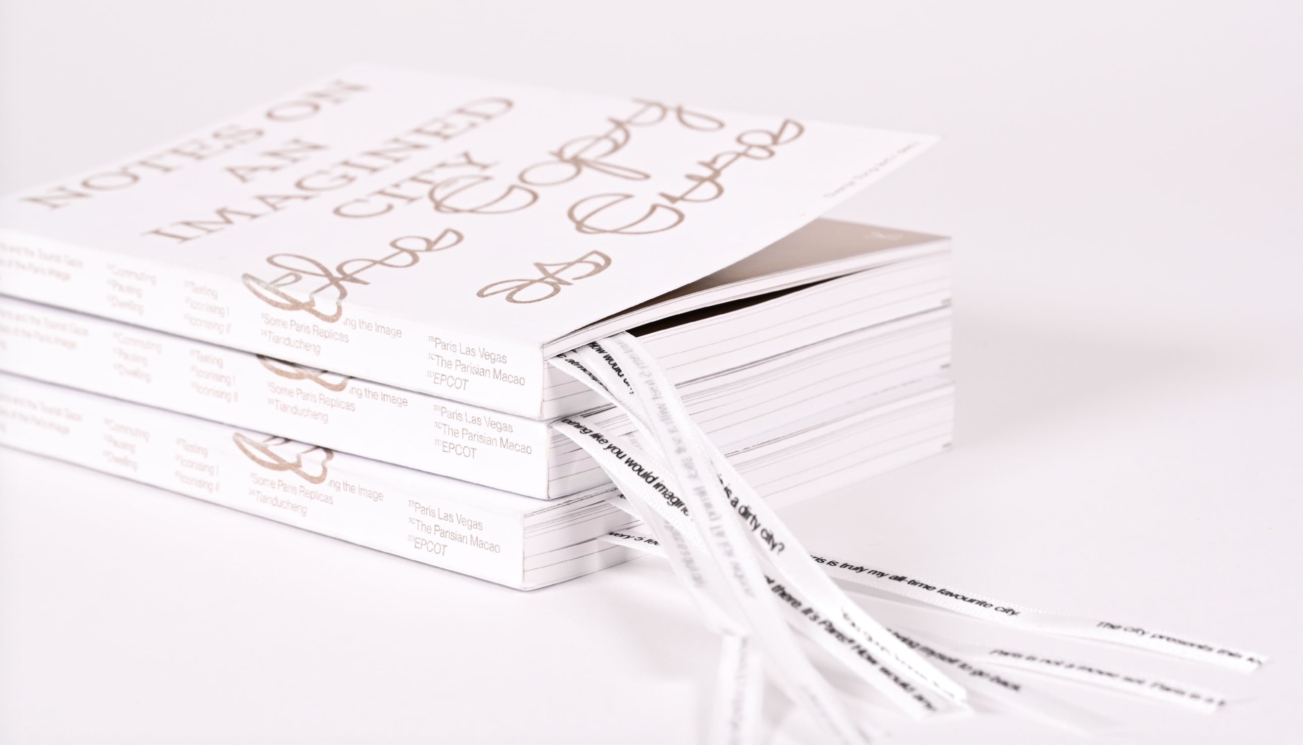 Three stacked copies of a cream-coloured bound book with elegant title 'Notes on an Imagined City'. Fabric ribbons are extending out from the bottom of the books with black text on them.