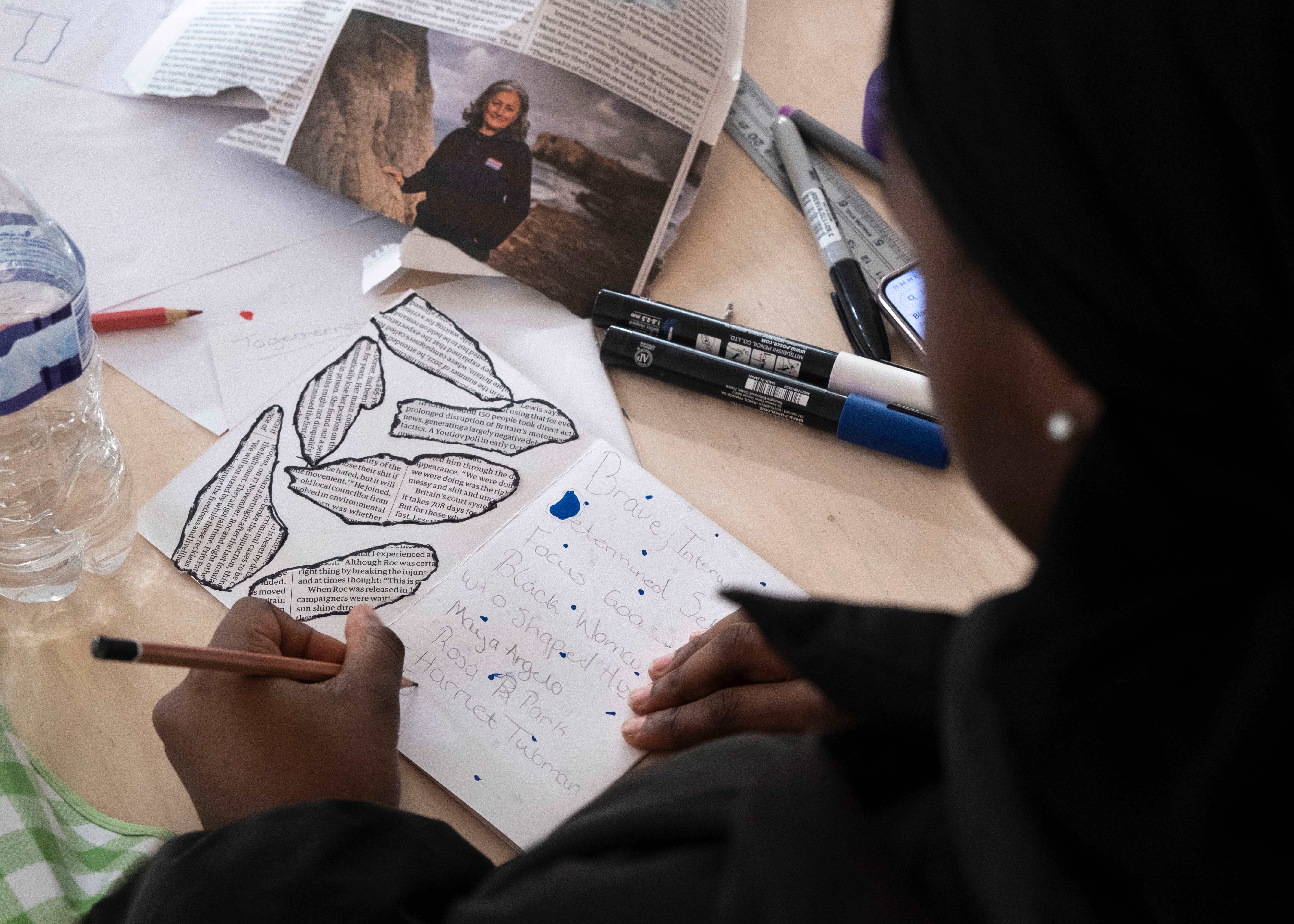 Person in black clothing writes notes beside outlined leaf shapes on paper, with markers, a newspaper, and water bottle on the table.