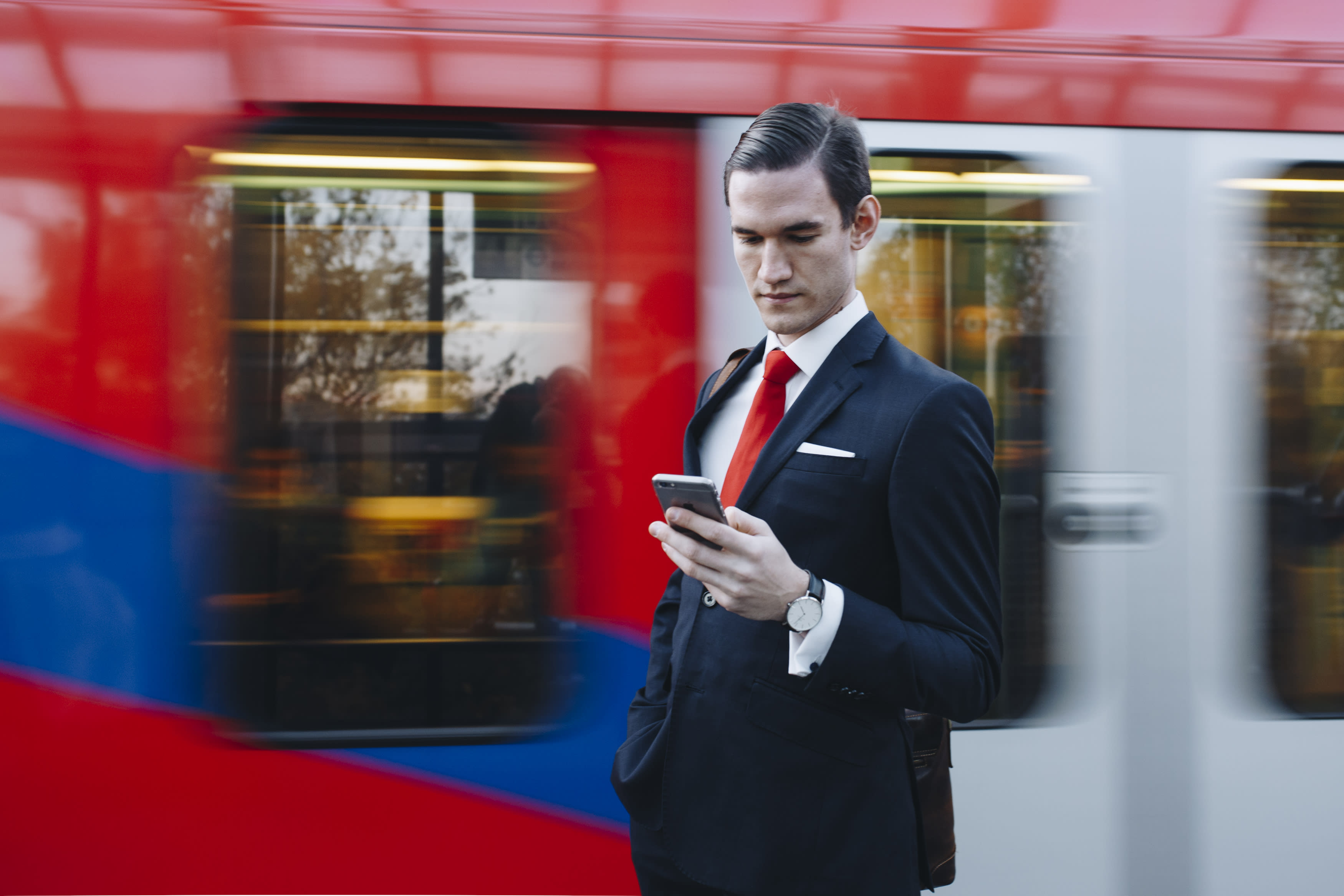 A person in a navy blue suit and a red tie standing in front of a passing train, looking into their phone.