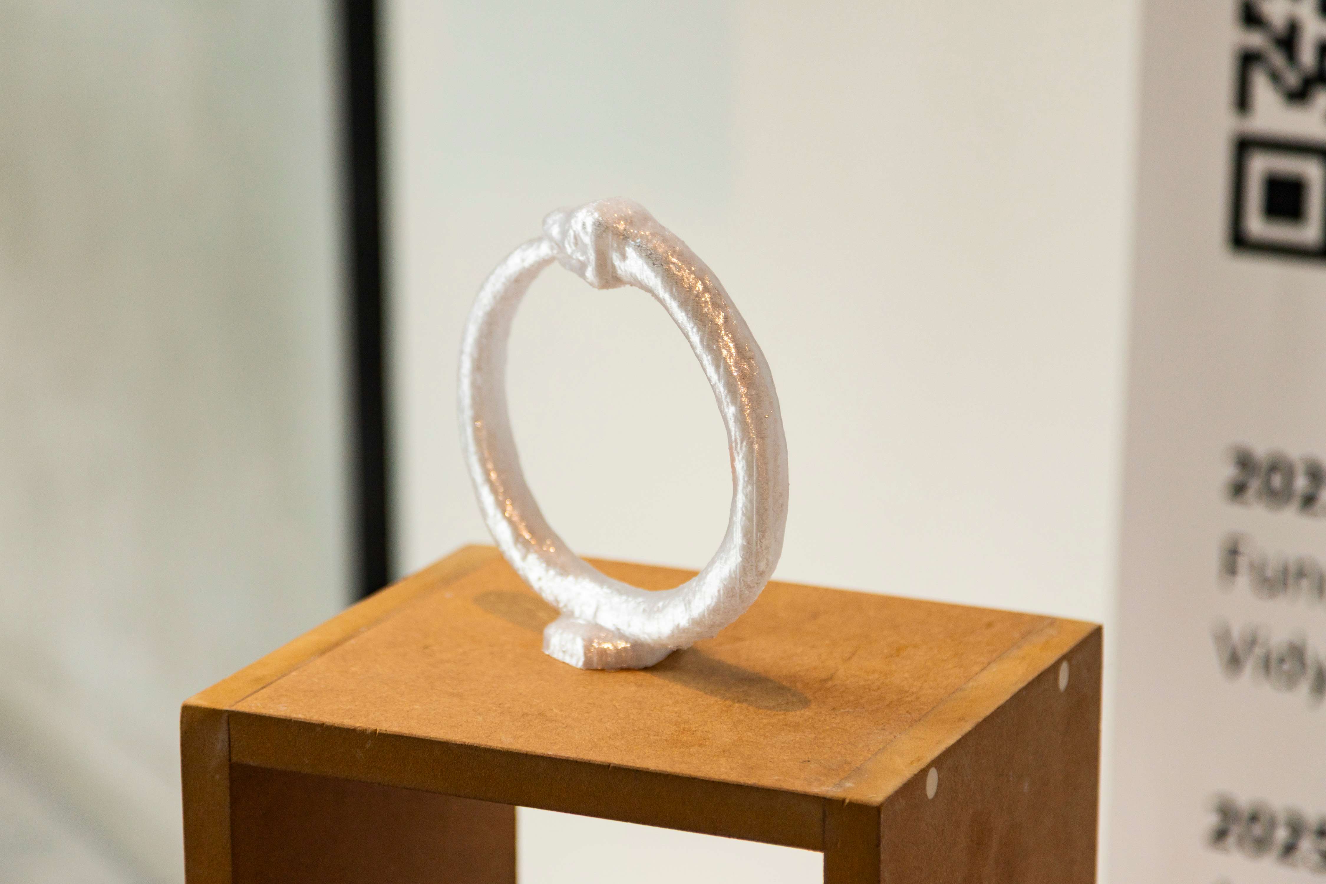 An upright silver piece of jewellery in the shape of an oruborous, a snake eating its own tail. It is placed on a wooden stand, against a white background.