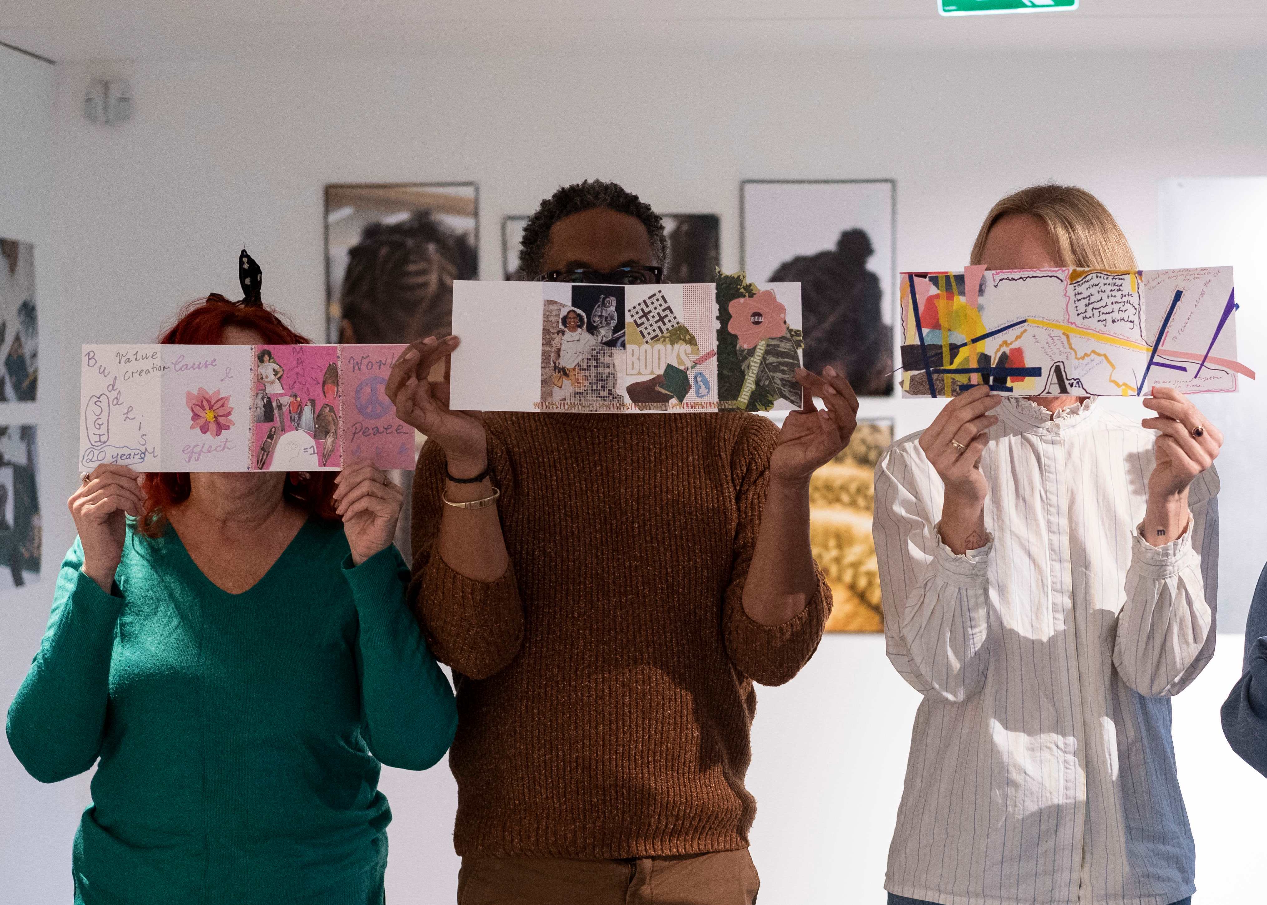 Three people in an art gallery hold colorful handmade collage cards over their faces, creating a playful, creative scene.