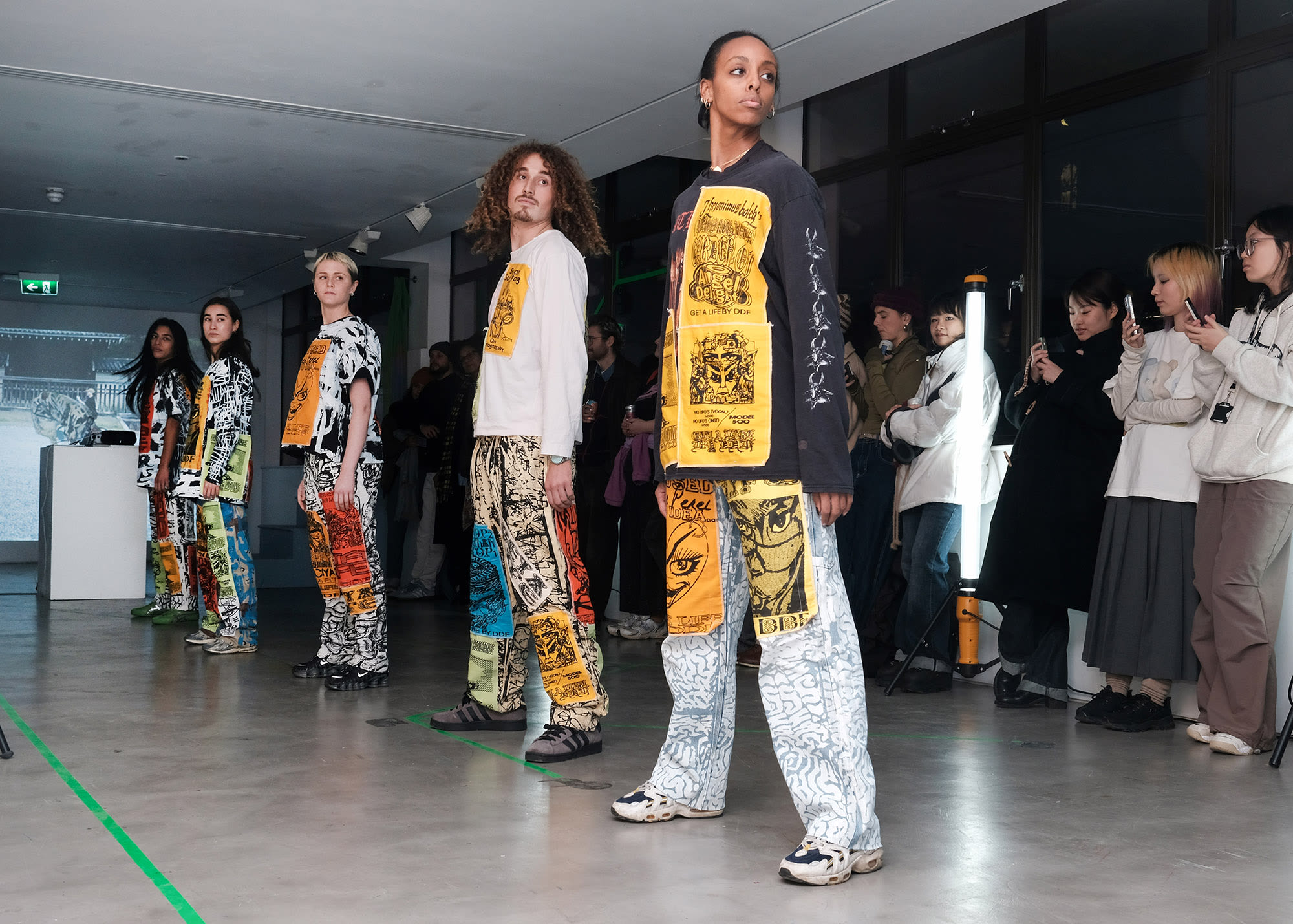 5 dancers standing in a row wearing clothes with large patches on that feature the artwork of Daniel David Freeman. The performance took place during the GET A LIFE exhibition at Camberwell Space.