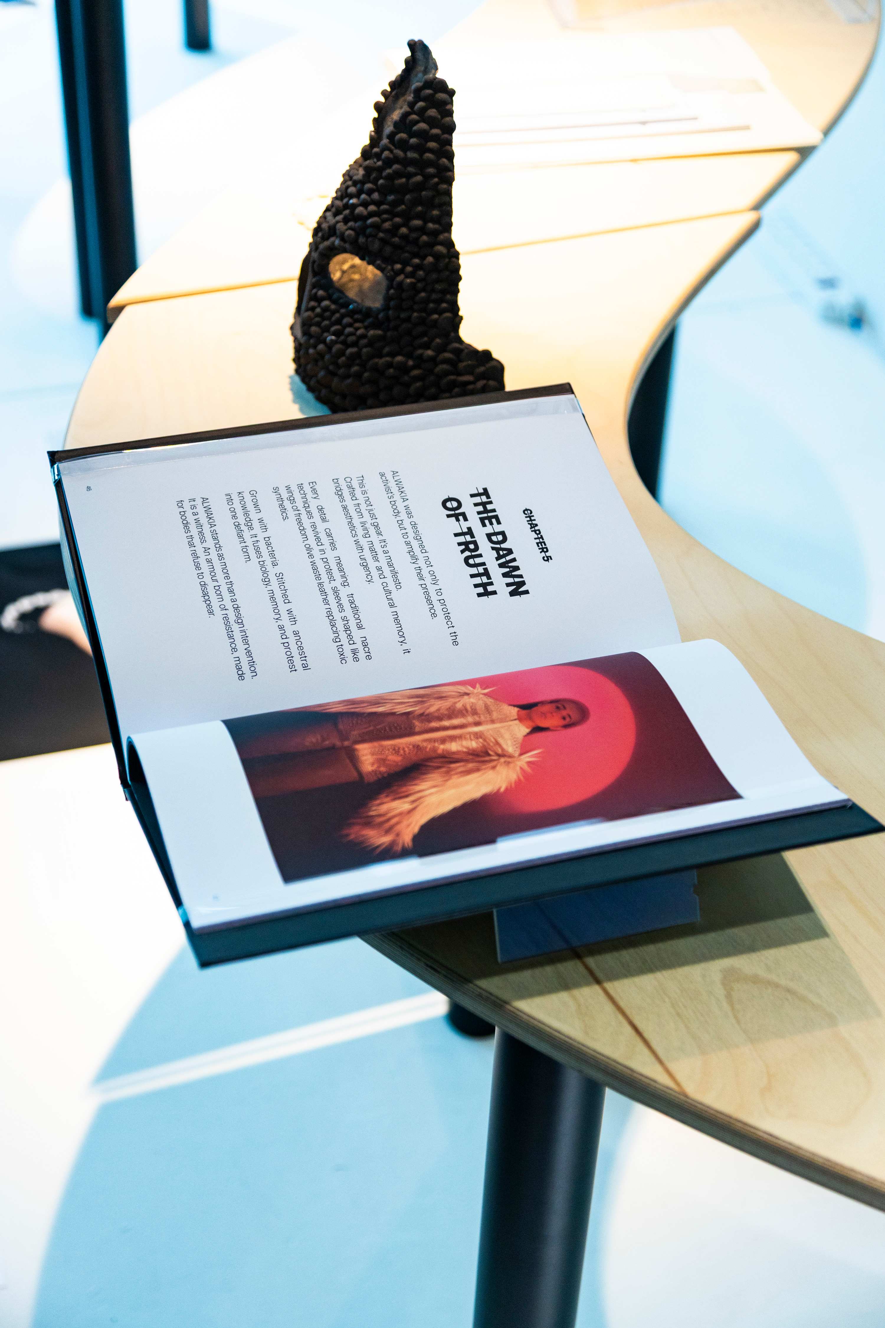 A publication is open on a page spread showing text on one side and an image of a feathered garment worn on a person on the other page. The book is placed on a curving table and there is a furry object standing to the right of it.