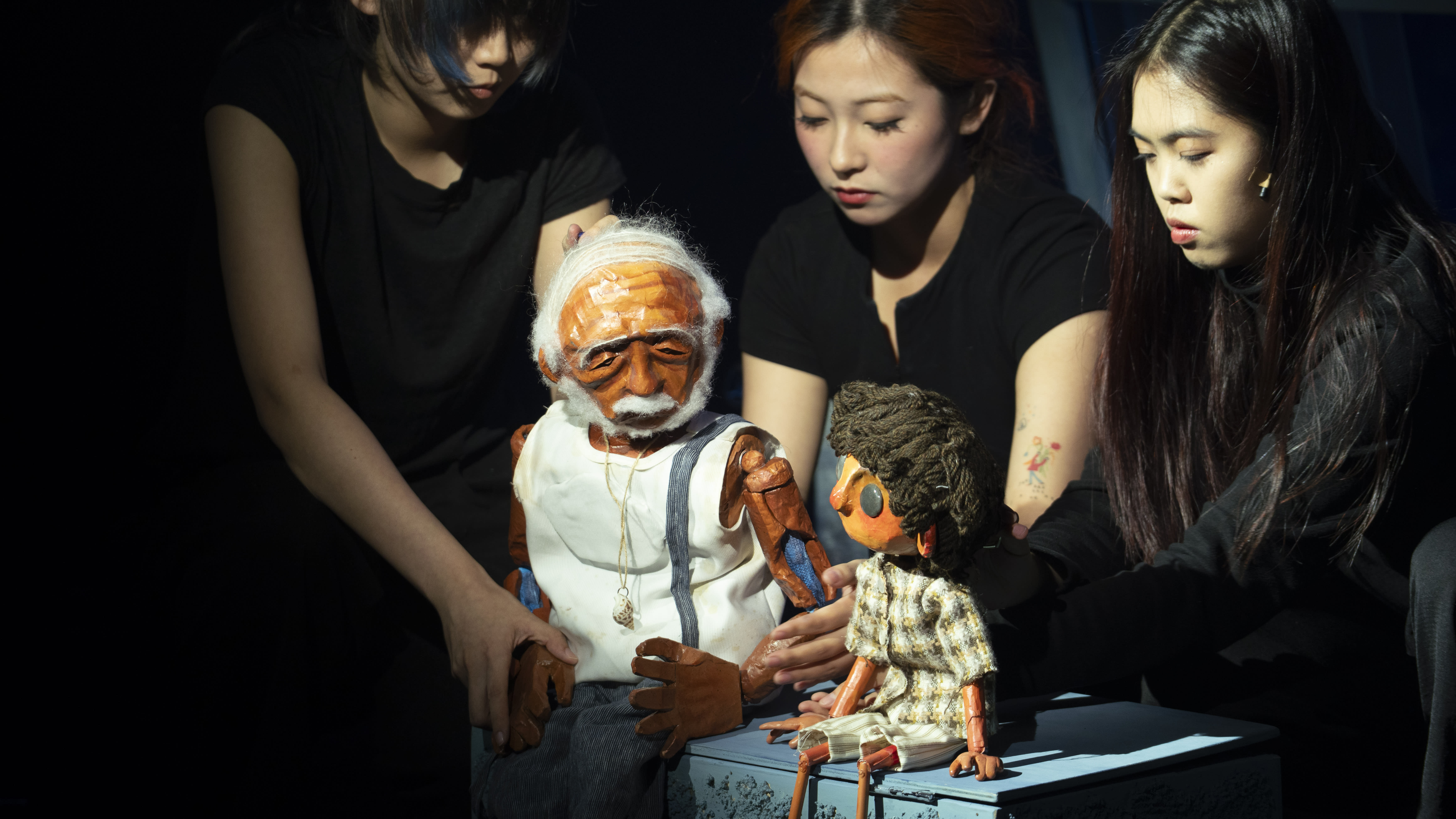 Three people manipulate two puppets onstage. One is an elderly man, the other a child. The scene is intimate, with a focus on storytelling.