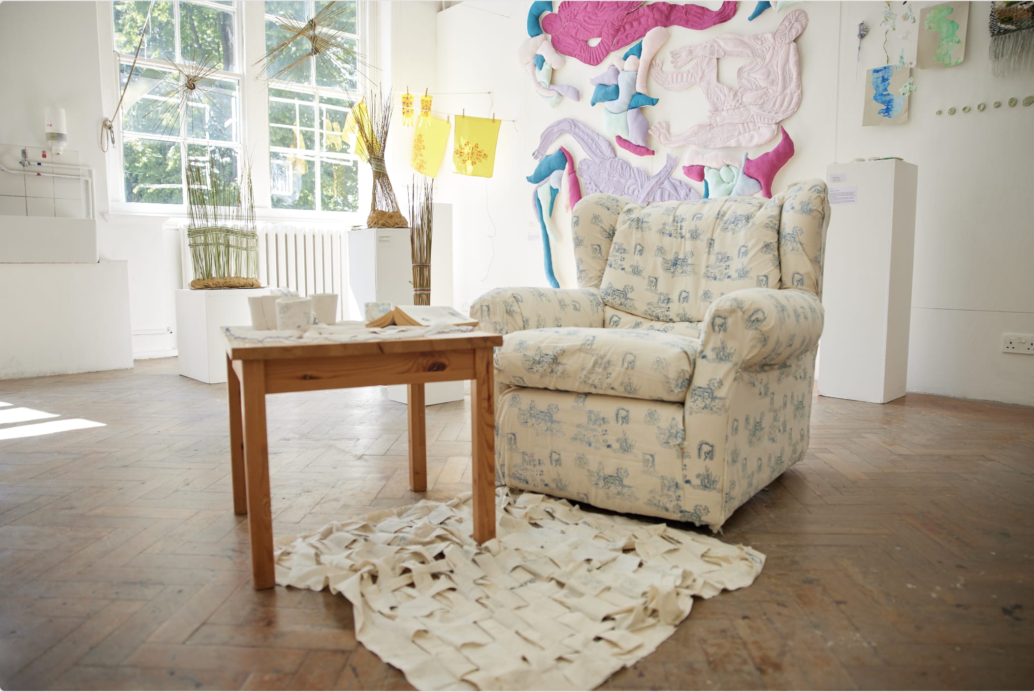 A white armchair upholstered with a soft blue toile de jouy fabric placed alongside a wooden table with various artefacts. There is a loosely woven rug underneath the table.