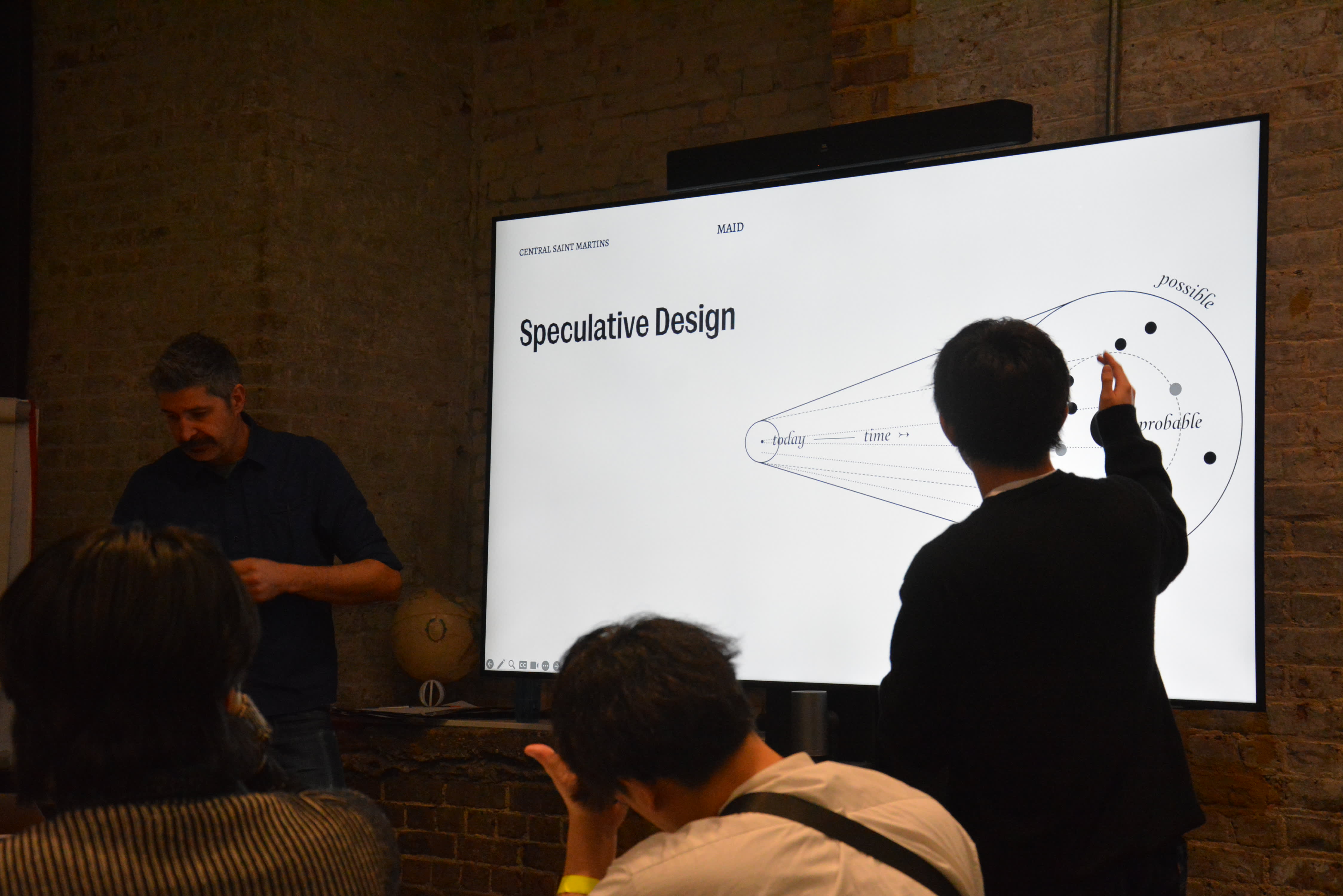 An image of two people in front of a screen with the words 'speculative design' on it